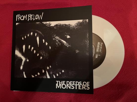 An album cover featuring the title "From Below" and "The Deeds of Monsters," with a dark, abstract illustration of a creature's open mouth displaying sharp teeth. A vinyl record in white is partially visible alongside the cover.