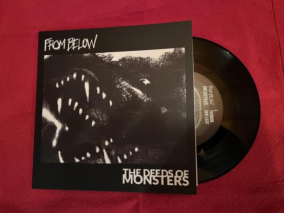 An image of a vinyl record with its cover partially open. The cover features dark artwork depicting a monstrous creature with sharp teeth and the text "FROM BELOW" and "THE DEEDS OF MONSTERS."