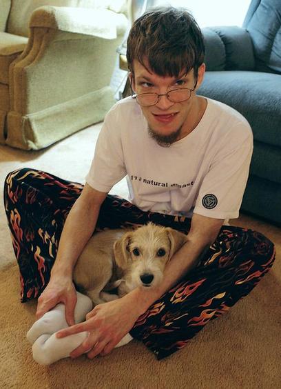 Young man sitting on floor cross legged with very young puppy Buddy cuddled in his lap. Man is smacking and Buddy looks like he is not going anywhere soon.