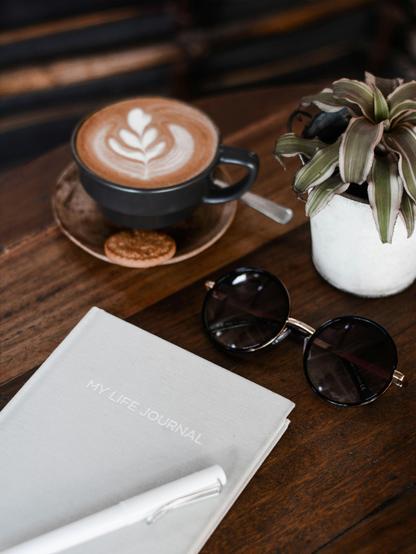 black ceramic mug with latte art sits on a small saucer, alongside a small cookie. A small white pot containing a green and white striped plant is also present. A pair of black sunglasses rests on a lightly textured, open notebook with the text “MY LIFE JOURNAL” printed on the cover in capital letters. The lighting appears soft, casting subtle shadows and highlighting the textures of the objects.
