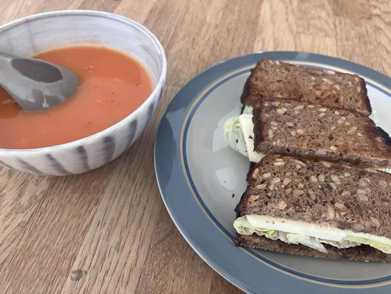 A bowl of tomato soup with a spoon beside it, accompanied by a plate featuring three slices of brown bread filled with lettuce and cheese. The setting is on a wooden surface.