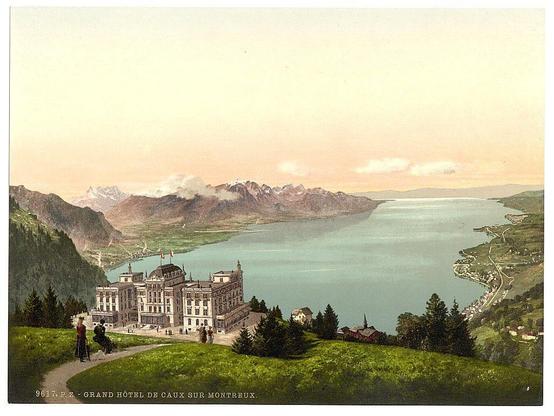 The image is a vintage hand-colored lithograph depicting the Grand Hotel de Caux, located on the shores of Lake Geneva in Switzerland. It shows an expansive view from atop a lush green hillside overlooking the hotel and its surroundings. The architectural style of the building suggests it was constructed during or shortly after 1900.

The scene is serene with a clear sky that hints at either sunrise or sunset, as indicated by soft pink hues near the horizon which gradually fade into lighter blue tones above. A calm lake reflects these colors on its surface and extends to meet majestic snow-capped mountains in the distance under an expansive sky.

In front of the hotel, several individuals are seen: two people appear conversing beside a path leading towards the building's entrance, while others sit or stand near various points around what seems like balconies or windows. The environment is picturesque with verdant trees and manicured lawns enhancing the idyllic setting.

A winding road snakes down from higher ground to meet another roadway along which houses are placed in close proximity to each other, showcasing a blend of residential architecture within this tranquil Swiss locale.