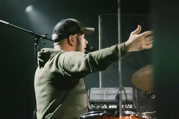 Ein Mann in Kapuze singt in ein Mikrofon und spielt Schlagzeug. Er trägt eine Kappe und hat ein In-Ear-Monitor im Ohr. Im Hintergrund ist ein Scheinwerfer und weiteres Equipment zu sehen.
