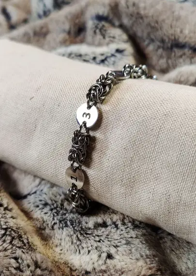 Bracelet en maille byzantine, dont les modules sont séparés à 4 reprises par des chiffres : 1312. Montré ici sur un présentoir tubulaire pour lui donner la même forme que lorsqu'il est porté.
