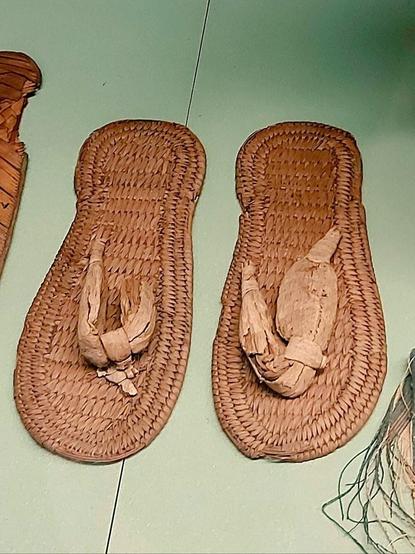 The picture shows a completely preserved pair of Egyptian sandals, they resemble modern flip flops. They are made of woven reed and palm leaves, the sole with bound edges.