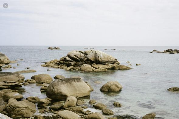 Felsige Küstenlandschaft mit großen, rund geschliffenen Granitblöcken im flachen, klaren Meer; ruhige Wellen unter hellem, leicht bewölktem Himmel.
