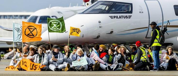 Menschen mit Extinction Rebellion Fahnen sitzen vor Flugzeugen auf einem Flugplatz