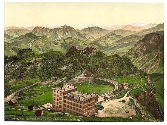 The image shows a picturesque landscape dominated by lush green mountains and valleys, with several structures nestled within the terrain. In the foreground is an expansive stone building on elevated grounds, featuring numerous windows and surrounded by smaller outbuildings or possibly cottages. A winding road snakes around to connect different parts of this complex.

Behind it lies rolling hills dotted with trees, indicative of a high-altitude environment. The background reveals majestic mountain ranges under a bright sky tinged with hues suggesting either dawn or dusk light. Snow-capped peaks add contrast and suggest the location's elevation.

The image appears as an illustration rather than a photograph, likely created using lithography techniques such as photochrom to reproduce colors from original photographs. This method was popular for travel publications in the late 19th century. The print bears text that indicates it is related to "Rochers de Naye," which translates to Rockerfellers' Peak or Rochers-de-Naye, a famous mountain near Geneva Lake in Switzerland.

The overall impression conveyed by this image is one of serene beauty and historical allure, capturing the essence of Swiss alpine scenery during its period as an accessible destination for European visitors.