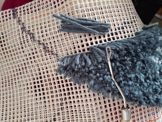 Ein Detailfoto von einem angefangenen Knüpfteppich. Auf großmaschigem Stramin-Gewebe ist ein Kuchenstück-förmiger Bereich mit grauer Wolle beknüpft. Auf dem Stramin liegen mehrere 6cm lange Wollstücke bereit und ebenso der Knüpfhaken.