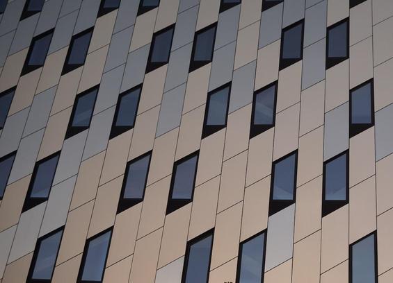 A facade of a building is flat and smooth, made of parallelograms that are either blue or beige. The windows are also parallelograms and arearranged in loose hexagons.
There appears to be a graduated tint, brighter in the lower right and darker in the top left, but this is an effect of the light and the arrangement of blue and beige panels. This is a photograph, not a computer render.