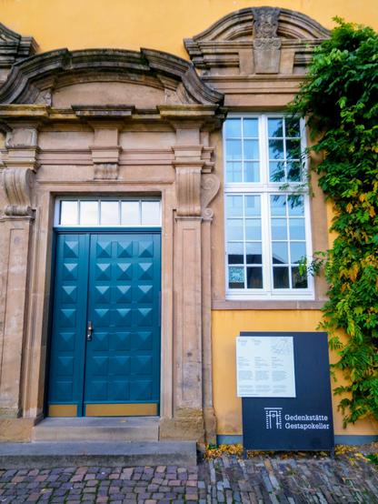 Eingang im Westflügel des Osnabrücker Schlosses: eine blaue Tür mit portalartiger Umrahmung aus Naturstein in gelber Fassade, daneben ein hohes Sprossenfenster mit Fensteraufsatz ebenfalls aus Naturstein unter dem das Hinweisschild der Gedenkstätte Gestapokeller steht.