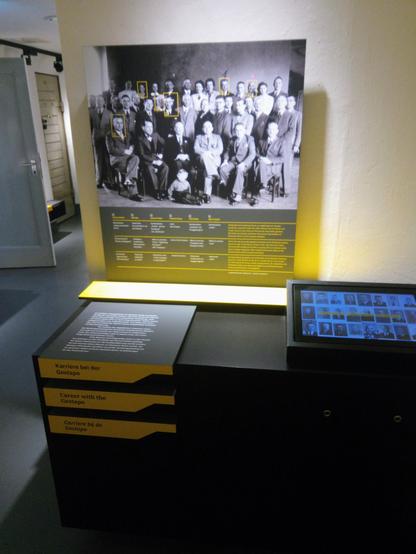 Ausstellungsstation "Karriere bei der Gestapo" mit einem an der Wand groß aufgezogenen Gruppenfoto, das Angehörige der Gestapo Osnabrück 1940 oder 1941 zeigt (Männer und Frauen in Zivilkleidung in mehreren Reihen sitzend und stehend), darunter ein schwarz-gelbes Ausstellungsmöbel mit Erläuterungstext links und rechts einer Medienstation zum Abrufen von biografischen Informationen zu den Gestapo-Leuten.
