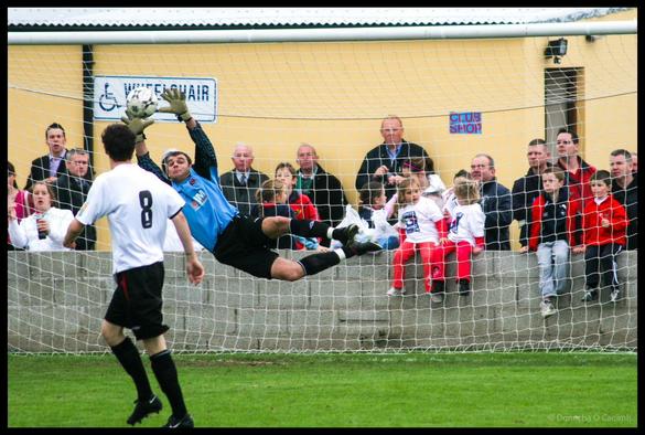 Dramatic action shot of goalkeeper in blue jersey diving horizontally through air with arms outstretched attempting to save shot during local football match, with player number 8 in white and black visible in foreground and crowd of spectators including children in red and white jerseys watching from behind goal net at community football pitch with yellow corrugated wall and wheelchair sign visible in background.