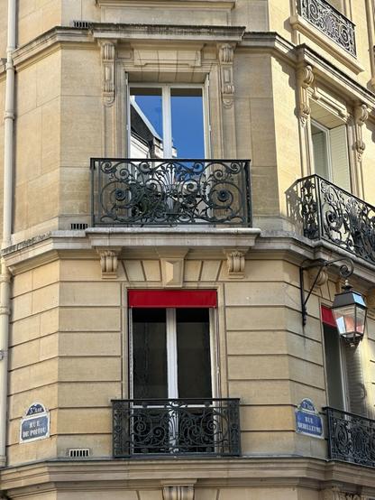 two floors of floor-height windows on the corner of a Parisian apartment building, with shallow, ornate wrought-iron balconies and red awnings. The upper window reflects the blue sky and an adjacent building