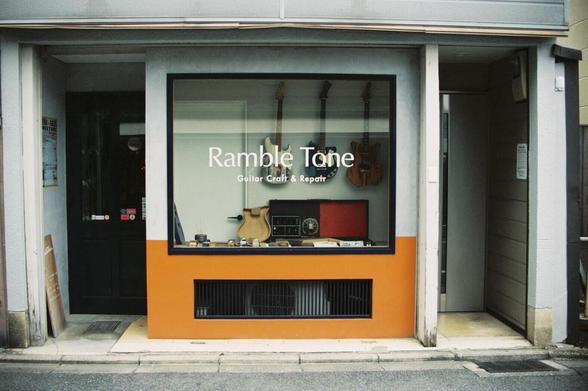 This image shows a storefront with a large window and an orange lower section. The window displays the text "Ramble Tone" in large white letters, with "Guitar Craft & Repair" in smaller white letters beneath it. Inside the window, several guitars (one light-colored, one dark, and one red) are mounted on the wall, along with what seem to be a red amplifier and small items on a shelf. To the left of the window is a black door; to the right is an open doorway. The building’s exterior is light gray, with a black window frame. The sidewalk and street are visible at the bottom of the image.