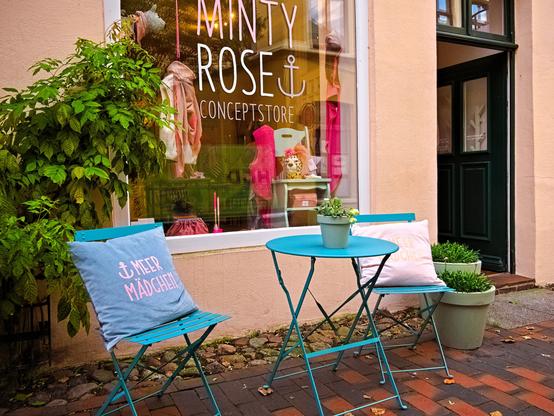 In front of a shop called ‘Minty Rose’ are two folding chairs and a table. They are painted in a pretty shade of blue. On each chair is a cushion with the words ‘Meer Mädchen' (Sea girl) written on it. The shop window is decorated with clothing and accessories, all in shades of pink and brown. This matches the front of the building, which is light in colour with a pinkish-brown tinge. Next to the shop window is an open green door. There are also plants in front of the window. I think it looks inviting.