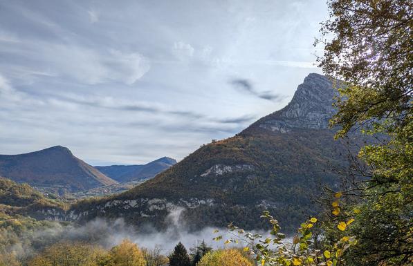 Behind a strand of mist in the valley, a rock-capped pointy mountain rises, and behind it in the distance two more. The leaves of many or the trees are turning gold and orange.