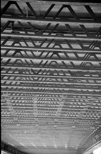 Das Schwarzweiß-Foto zeigt die Stahlstrebenkonstruktion unter der Autobahnbrücke über das Fechinger Tal. Die Streben gehen längs und sind nach oben durch schräge doppelt V-förmige Streben mit der Brückendecke verbunden. Durch die Perspektive sieht es so aus, als würden sie nach unten hin immer näher aneinander rücken.