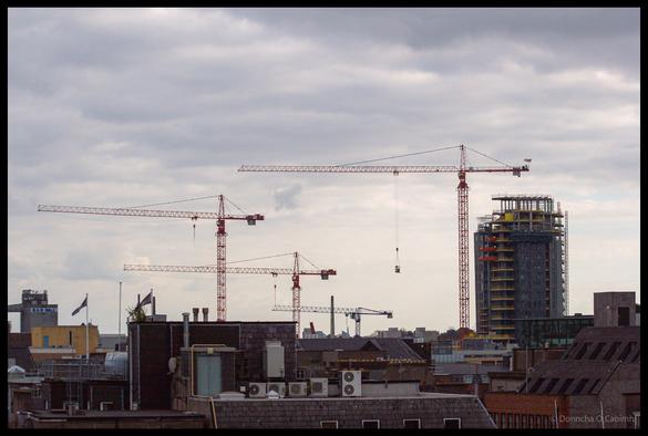 Urban skyline view of Cork city rooftops in 2007 showing four tall construction cranes dominating cloudy grey sky, with the Elysian tower under construction on right side, period buildings with satellite dishes and air conditioning units visible in foreground, capturing Cork during its Celtic Tiger era building boom.