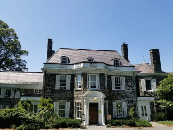 The image shows a large, ornate building with a stone facade, multiple chimneys, and a symmetrical design. The building has several windows and an entrance with a decorative arch and a yellow circular feature above it. The surrounding area has greenery and trees, set against a clear blue sky.

Image Credits: Wikimedia / Antigng / CC BY-SA 4.0