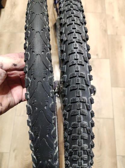 Dwie rowerowe opony obok siebie. Po lewej trochę węższa opona z gładką centralną częścią i małymi wypustkami po bokach. Po prawej trochę szersza opona z dużymi klockami bieżnika na całej szerokości.