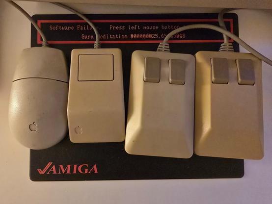 2 Classic Macintosh mice and Two Commodore (Amiga) Tank mice side by side on top of an Amiga GURU meditation mouse pad