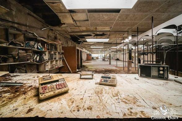 Three old calculators and a vintage radio sit on a dirty counter in an abandoned, dilapidated building with damaged ceiling tiles and empty shelves.