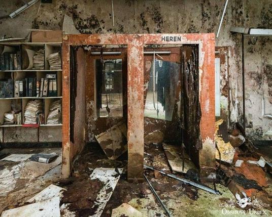 A decaying, abandoned office with a rusted, damaged booth labeled "HEREN," scattered debris, moldy walls, and disorganized shelves full of old files.