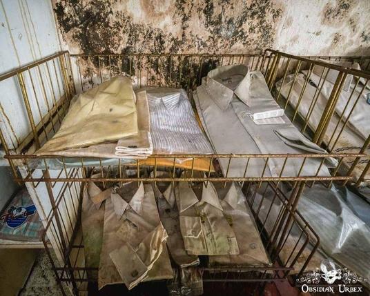 Neatly folded, aged shirts sit on rusty metal shelves in front of a stained, peeling wall in an abandoned building.