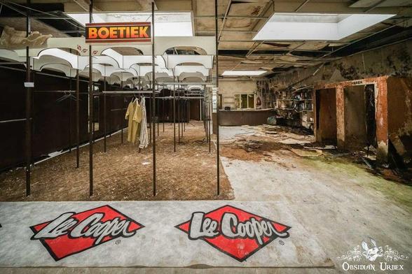 Abandoned clothing store with empty racks, damaged ceiling tiles, debris on the floor, and “Lee Cooper” and “Boetiek” signs visible.
