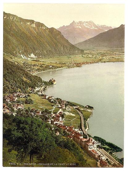 This image depicts a picturesque view from above, showcasing the landscape around Lake Geneva in Switzerland. The scene includes rolling hills dotted with small settlements and roads meandering through them. A large body of water is visible on the left side, likely representing part of Lake Geneva's shoreline. To its right are more elevated terrains leading up to imposing mountains. These mountain ranges have patches that appear to be covered by snow or frost, suggesting a high altitude.

The image has an aged look with faded colors and text at the bottom indicating it might be from a vintage postcard or print. Notable features include dense greenery covering parts of the hills, contrasting with cultivated fields in other areas. The town appears quaint and peaceful, nestled between natural beauty and human habitation. A railway track runs parallel to one side of the lake, hinting at accessibility for travel.

The photo's caption identifies it as "Chillon et la Dent du Midi," indicating a connection to notable Swiss landmarks like Chillon Castle on Lake Geneva and possibly Montbodif (also known as Dent du Midi), which is a mountain peak in the Jura mountains. The overall impression of this image is one of serene beauty, historical charm, and natural splendor characteristic of Switzerland's Alpine region during late 19th to early 20th century times.
