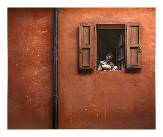 A person with short, dark hair is wearing a beige zip-up and is sitting inside of a small, rectangular window. It has open, weathered brown wooden shutters. The wall is textured and painted in a warm, rusty orange color. A black drainpipe runs vertically on the left side. The window is near the right hand corner of the photo.