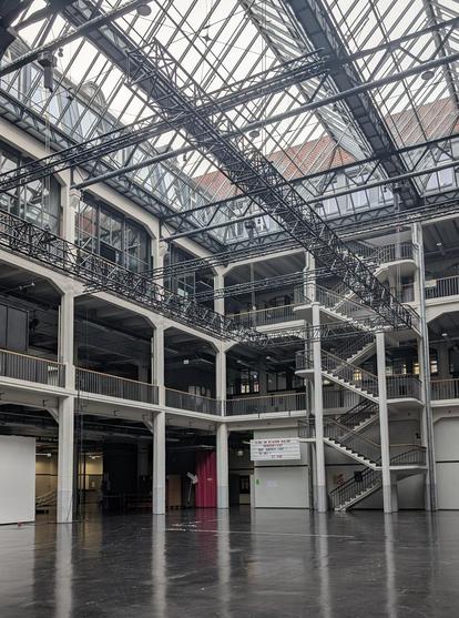 einer der Lichthöfe der HfG Karlsruhe. man sieht einen leeren Raum, der auf drei Geschossen von einer Galerie umarmt wird. Oben hängen Konstruktionen die zur Befestigung von Technik gedacht sind