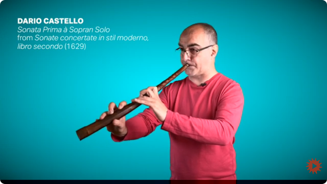A musician in a salmon shirt, Dario Castello, plays a long, thin, horn-shaped instrument called a cornett.