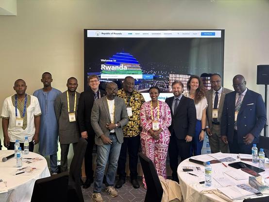 A group of twelve people, including men and women in business and traditional attire, stand together and smile for a photo in front of a large screen displaying Welcome to Rwanda at a conference event.