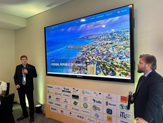 Two men in suits stand in front of a large screen displaying a website for the Federal Republic of Somalia, featuring a coastal city view. The foreground shows a wall with multiple sponsor logos.