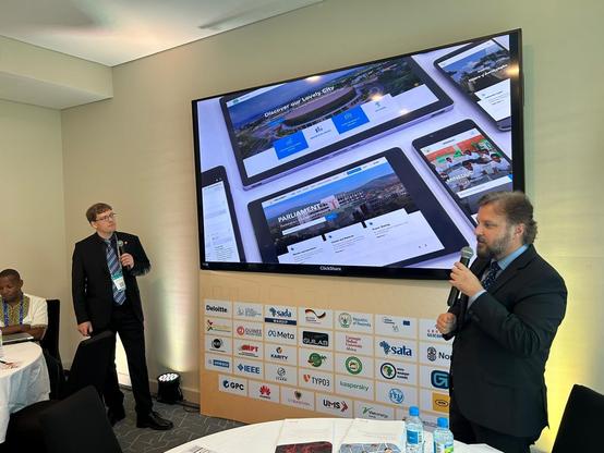 Two men in suits stand and speak with microphones in front of a large screen displaying website designs on digital devices at a conference. Other people sit at round tables, and sponsor logos are visible on the stage.