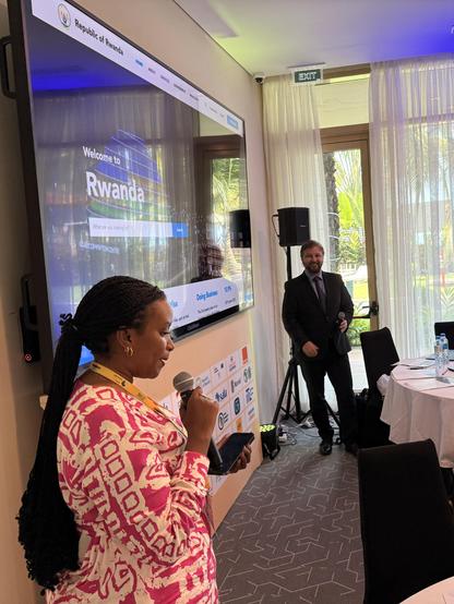 A woman speaks into a microphone in a conference room, with a man standing near a screen displaying Welcome to Rwanda and daylight visible through sheer curtains in the background.