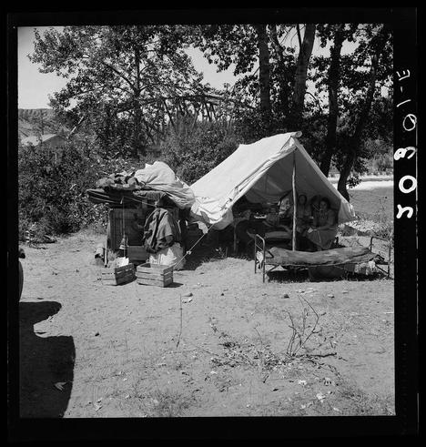 The image depicts a makeshift campsite with several individuals gathered under and around a large tent. The setting appears to be an outdoor area, possibly in the countryside or rural environment. Various personal belongings are scattered around: blankets on top of what seems like cots for sleeping, bags containing supplies or clothing stacked beside wooden crates holding items that could include foodstuffs, utensils, or other necessities.

A notable feature is a folding chair with someone seated within it; however, their identity remains indistinct. The area surrounding the campsite features trees and shrubbery indicative of an uncultivated landscape. A vehicle's tail end can be seen on the leftmost side of the frame, suggesting that this might have been the family's mode of transportation.

The image is rendered in black and white, which adds a historical or documentary quality to it. The caption provided indicates that this campsite belongs to a migratory Texas family who are currently residing at Ramblers Park in Yakima Valley, Washington as documented by Dorothea Lange around 1938-1940 during the Great Depression era.

The image serves not only as an illustrative representation of temporary living conditions but also underscores the challenges faced by families displaced due to economic hardships or migratory patterns.