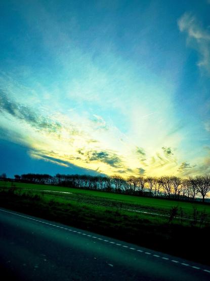 A scenic view features a blue sky with streaks of clouds and a golden horizon. A row of trees is silhouetted against a green field, alongside a road in the foreground.