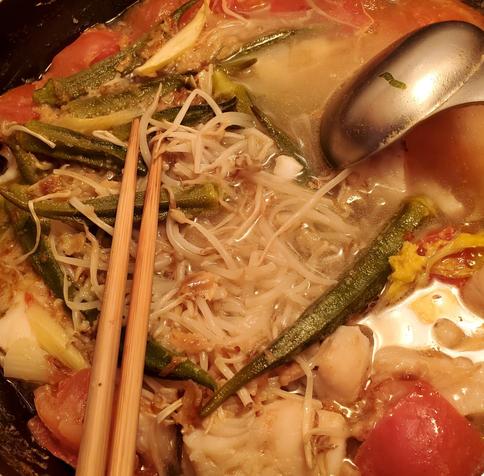 Close-up shot of a pot of canh chua ca soup, featuring okra, tomatoes, bean sprouts, and cod fish in a broth, with chopsticks laid on the side. A metal ladle is visible in the wok. The soup is in a big black wok.
