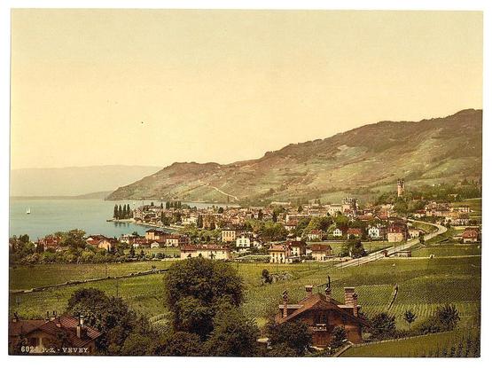 The image is a vintage postcard depicting the scenic town of Vevey on Lake Geneva in Switzerland. It showcases an expansive view with rolling hills and vineyards stretching into the foreground, indicative of viticulture prevalent in this region. The village itself features traditional European architecture with red-tiled roofs and varying building heights that create a picturesque skyline along the lake's edge.

In the background, Mount Salève dominates the landscape with patches of greenery on its slopes, suggesting it is either late spring or early summer when vegetation begins to flourish after winter dormancy. The calm waters of Lake Geneva reflect some of this scenery while boats are faintly visible, hinting at leisure activities in the area.

The postcard bears a caption "Vevey" and an artist's signature that reads "Léon L'Hermitte," indicating it was created by this particular Swiss photographer known for his photographic prints. The coloration is sepia-toned with hints of greenery, typical of early 20th-century print techniques.

Overall, the image captures a harmonious blend between nature and human settlement in Vevey during its postcard era, showcasing both economic activity (wine production) and leisurely pursuits against an idyllic backdrop.
