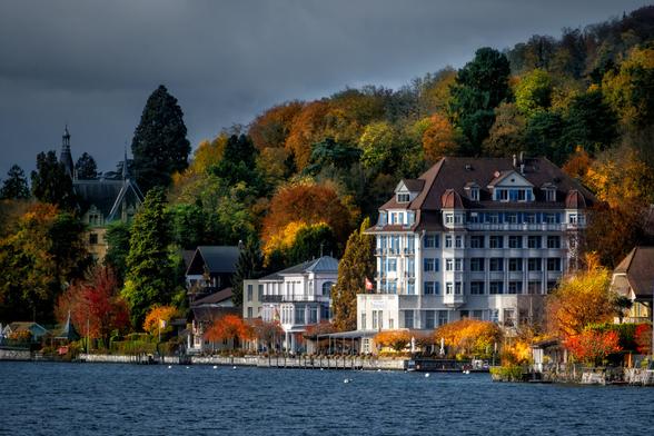 Eine herbstliche Szene am Ufer eines Sees, dominiert von einem grossen, weissen Hotelgebäude im Stil der Jahrhundertwende mit einem markanten braunen Walmdach. Die Fassade des Hotels, die viele blaue Fensterläden aufweist, steht im Kontrast zu den leuchtenden Herbstfarben der Bäume im Hintergrund. Ein dichter Wald mit Laub in Gold-, Orange- und Rottönen zieht sich den Hang hinauf unter einem dunklen, bewölkten Himmel. Links neben dem Hotel stehen weitere Villen und Häuser, darunter ein dunkelgrünes historisches Gebäude mit Turm, das fast vom üppigen Baumbestand verdeckt wird. Die Bebauung erstreckt sich entlang einer Uferpromenade mit Bootsstegen. Das dunkle Wasser des Sees, auf dem kleine Wellen sichtbar sind, nimmt den unteren Bildrand ein.