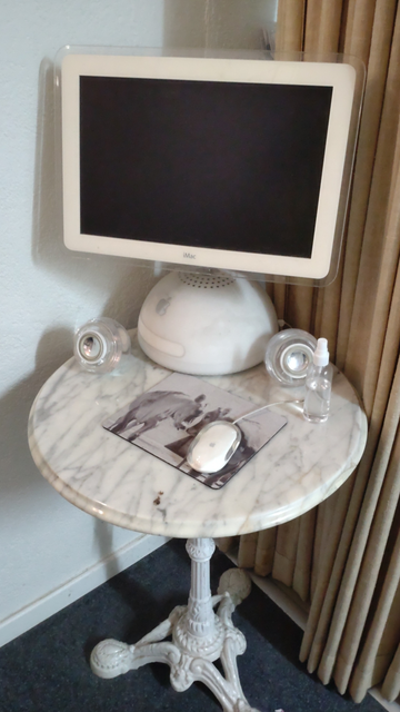 Ein vergilbter Nachttischlampen iMac steht auf einem kleinen Tisch mit Marmorplatte und Fuss Gusseisen. Dazu die originalen runden durchsichtigen Lautsprecher und eine weiss und durchsichtige Apple-Maus.