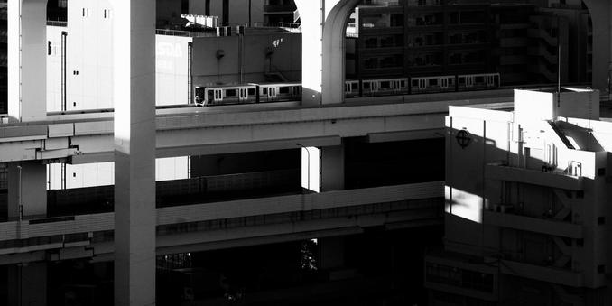 Black and white urban scene with elevated trains on intersecting tracks, surrounded by tall buildings. Strong shadows create a stark contrast against the structures, highlighting geometric patterns and architectural lines.