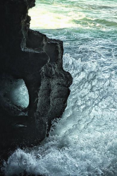 A vertical shot capturing a dramatic scene where a powerful wave crashes violently against a dark, rugged sea cave or cliff face. The rock dominates the left side, appearing intensely dark and textured, partially silhouetted. A small opening in the rock shows a turquoise glow. The right side is a brilliant, explosive plume of white and frothy water from the crashing wave. In the background, the sea is illuminated by a burst of sunlight at the upper edge, creating a bright contrast with the deep shadows of the foreground.