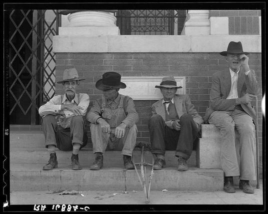 The image shows four men sitting on steps outside a brick building with classical architectural elements, such as corbelled columns and wrought iron fencing. They appear to be engaged in conversation or contemplation. From left to right: the first man is wearing overalls, suspenders, a wide-brimmed hat, eyeglasses, and has his arms folded; the second wears striped trousers with rolled-up sleeves, a dark top hat, and has one leg crossed over the other; the third sits on an elevated concrete block, clad in suit pants, dress shoes, and a fedora-style hat. The fourth man is dressed more formally than the others—suit jacket, tie, buttoned shirt—and holds his hand to his ear as if listening or speaking into something he’s holding up against it. A cane lies between their legs on the ground. There are no visible indicators of modern technology like phones; hence communication appears traditional rather than digital. The environment and attire suggest a setting from mid-20th-century America.