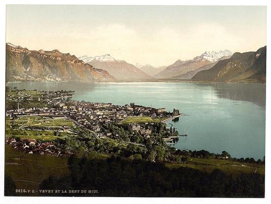 The image depicts a picturesque view of Geneva Lake, with the town of Vevey and Dent du Midi in Switzerland prominently featured. The vantage point appears to be high above the area, providing a sweeping perspective that encompasses both sides of the lake as well as its surroundings.

In the foreground on the right side, there's an expanse of lush green fields or vineyards stretching down towards the waterside town. The urban landscape includes clusters of buildings with varying architecture and sizes, suggesting residential areas along with possibly some commercial establishments closer to the waterfront.

The left shore is characterized by a denser concentration of structures, creating more defined streets and spaces among them compared to the right side's more open fields. This part also seems less developed than its counterpart on the opposite bank.

Beyond Vevey lies Dent du Midi in the background, with its rugged mountainous terrain extending into the distance where it meets distant snow-capped peaks. The sky above is a clear light blue, indicating fair weather conditions during the time of this photograph's creation. Overall, the image captures an idyllic and serene setting that illustrates both urban life by the lake and natural beauty surrounding Vevey in Switzerland around ca. 1890-1900.