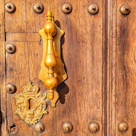 A close-up of a weathered, rustic wooden door dotted with round bronze nail heads. A large, ornate, highly polished golden brass door knocker is centered over a decorative escutcheon plate with a crown design.