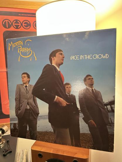 The image features a vinyl album cover titled "Face in the Crowd" by Merton Parkas. It shows four young men dressed in suits, standing on a beach with a pier in the background. The album name and band name are displayed prominently.